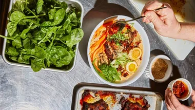 Un'immagine di piatti su un tavolo in acciaio inox, che comprende un vassoio con foglie verdi e una ciotola di ramen su cui viene versata una salsa rossa.