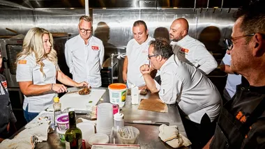 Un gruppo di chef di Unilever Food Solutions riunito attorno a un tavolo in acciaio inox mentre un collega prepara un piatto di pollo.